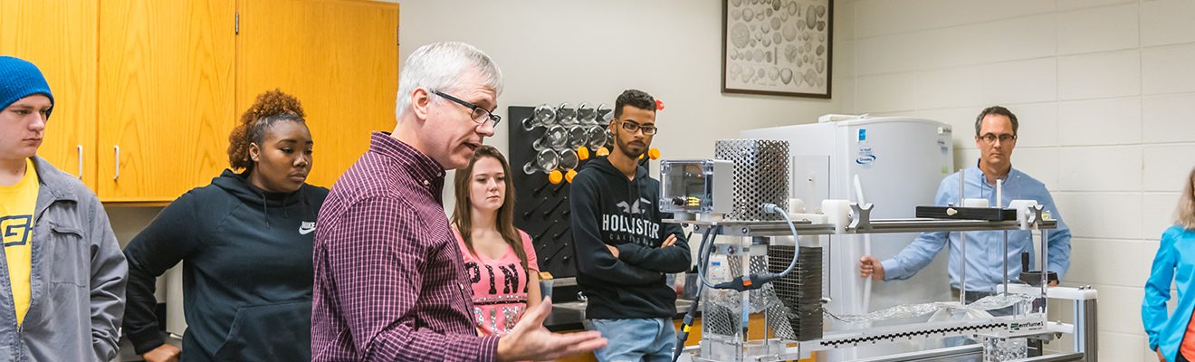 Geography professor standing in middle of science classroom teaches Geography degree students about a medium-sized geography apparatus on the table
