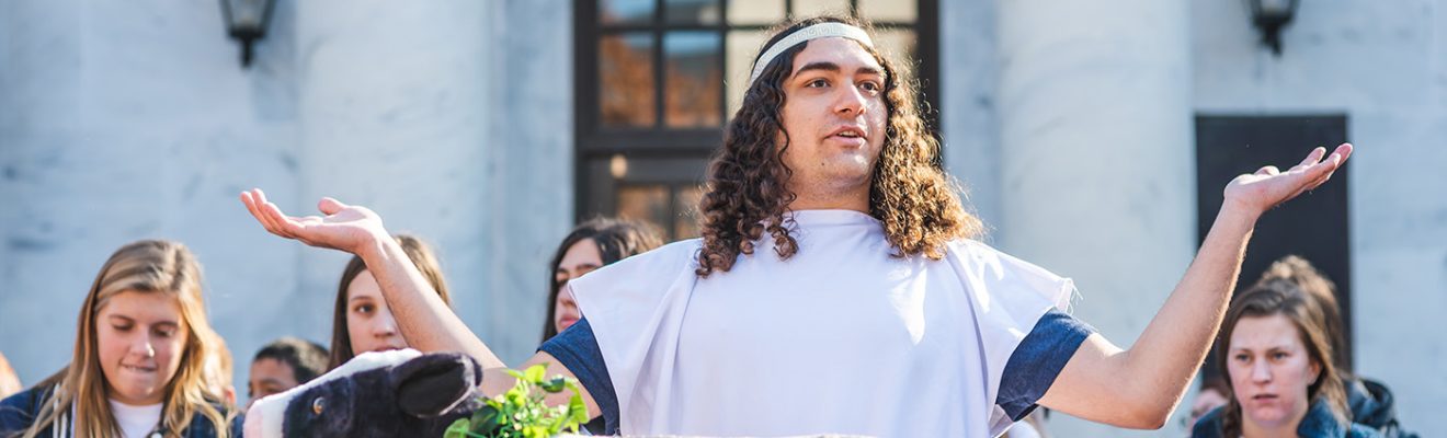 On Classics Day at UNC Greensboro, a man with his arms raised stands in front of a fake cow wearing greenery around its neck. In the background, a group of students watch