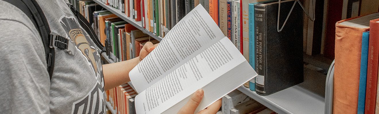 A female student wearing a UNCG t-shirt is reading a book while standing in the library