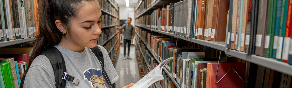 A young female student in a UNCG shirt is reading a book while standing in an aisle at the library