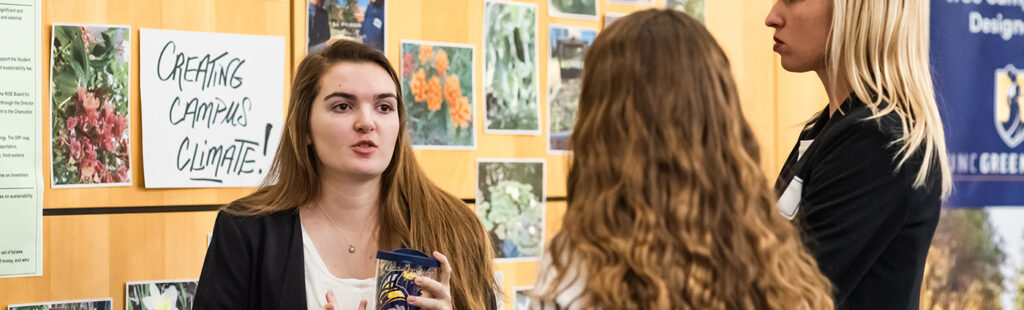 A Sustainability and Environment student is presenting a large wall filled with pictures of plants to two other students who are intently listening. The wall reads “Creating Campus Climate!”