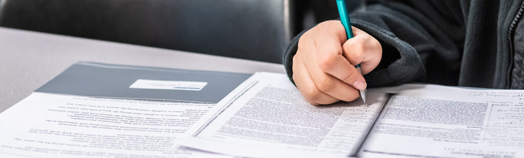 A close-up image of a hand holding a mechanical pencil, writing on sheets of paper filled with text.