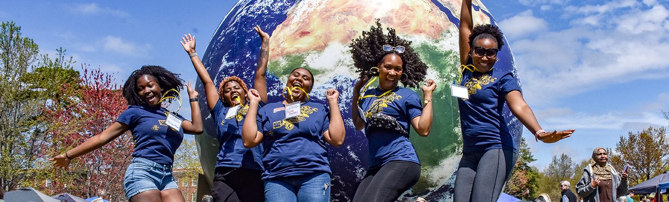 four women jump up in front of a massive model of the earth