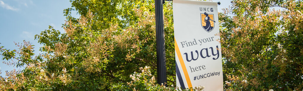 A light pole on UNCG’s campus surrounded by trees. On the pole is a banner with the UNCG Minerva logo and the words, “Find your way here. #UNCGWay”