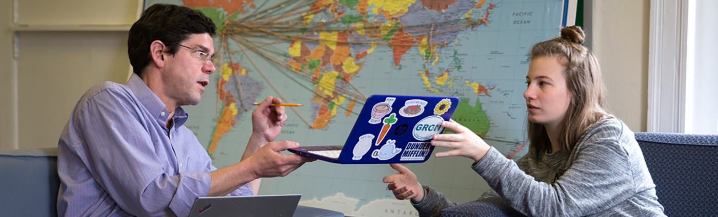 A professor and a student sit in arm chairs in front of a map of the world. The student holds a laptop out in front of the professor for their review.