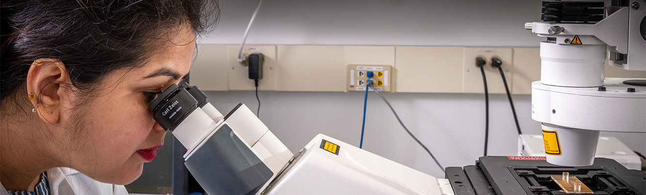A female student sits at a desk in a laboratory setting, she is peering through a sleek, state of the art magnification device.