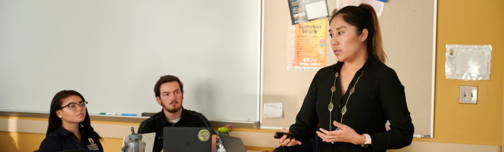 A Higher Education instructor, Dr. Delma Ramos, teaches in a classroom setting. Two students seated behind a desk are looking at the instructor.