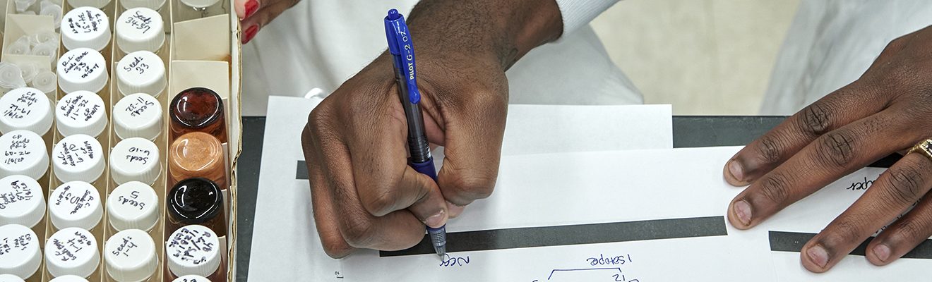 One set of hands writes notes, while another handles a collection of small bottles.