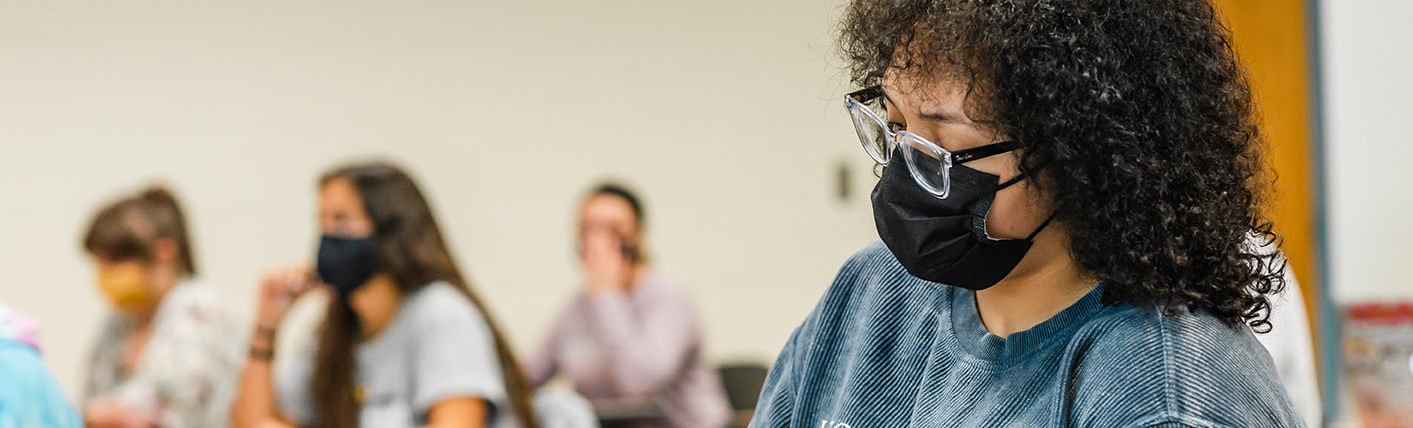 Female-presenting student sits at a desk in class taking notes. She is wearing a blue tie-dye UNC Greensboro sweatshirt.
