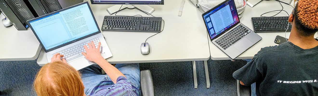 Two Geographic Information Science students work at desks in a computer lab with desktop computers and laptops to read and write information