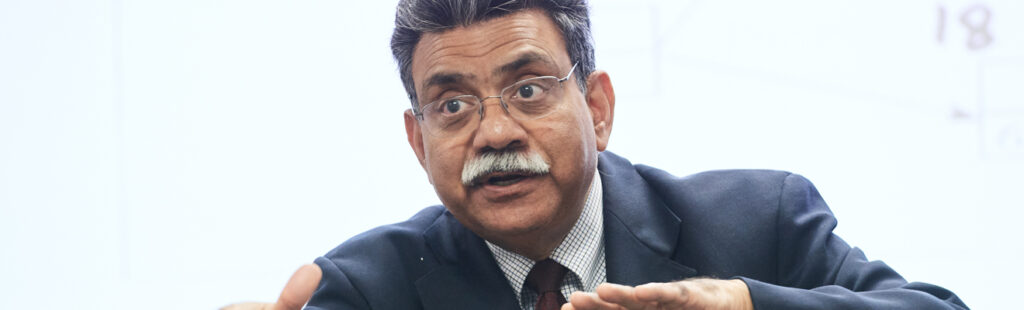 A UNCG Bryan PhD in Information Systems professor stands in front of the classroom. The photo is of him from the waist up. He is gesturing with both hands in an animated, playful manner as he lectures to the class.