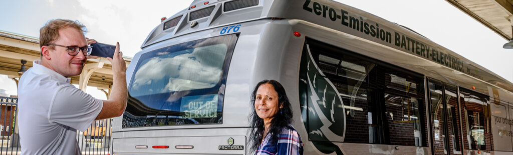 A male Urban Planning student and female professor take measurements with their phone at a bus station. Behind them is a zero admissions bus