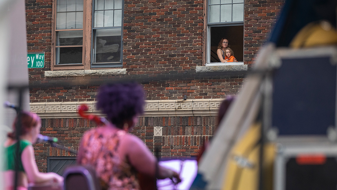 A mother and daughter watch singers at the 2022 NC Folk Festival from the window of their apartment.