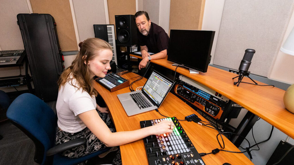 Professor leans over a table full of sound equipment, watching student working with an audio board and a laptop.