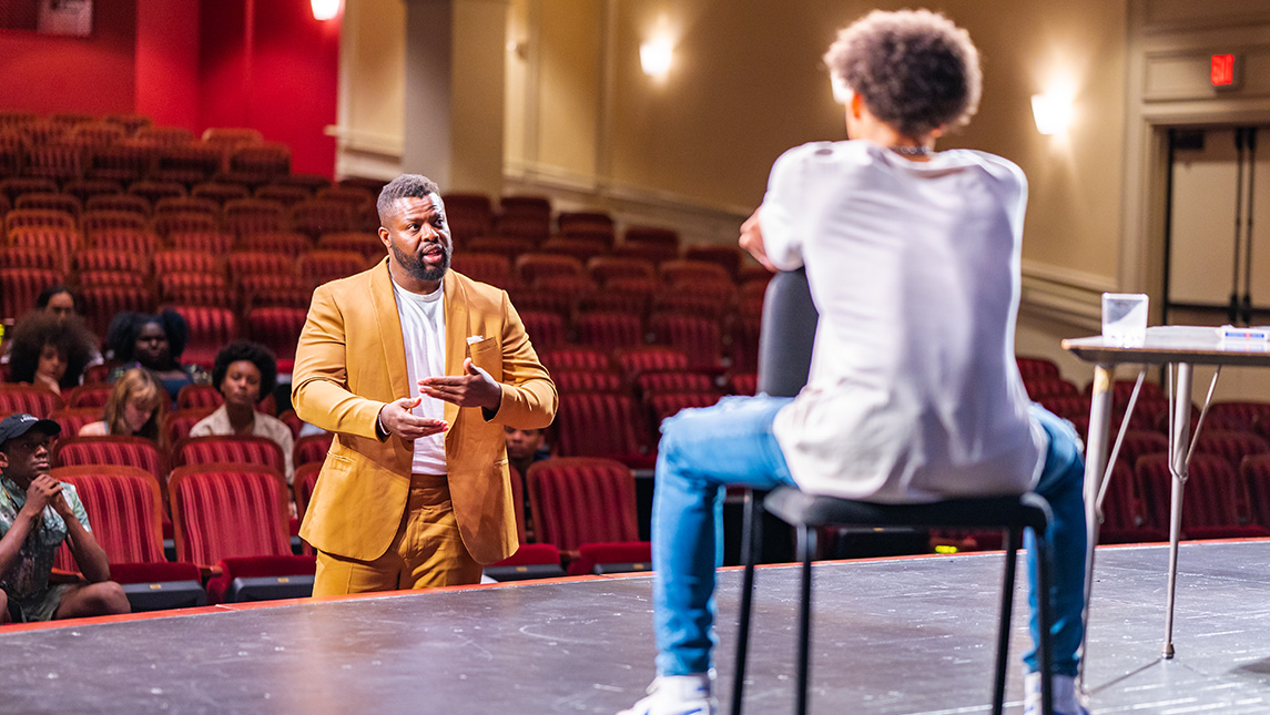 Actor Winston Duke stands in the auditorium gallery talking to student Xavier Henry seated on the stage.