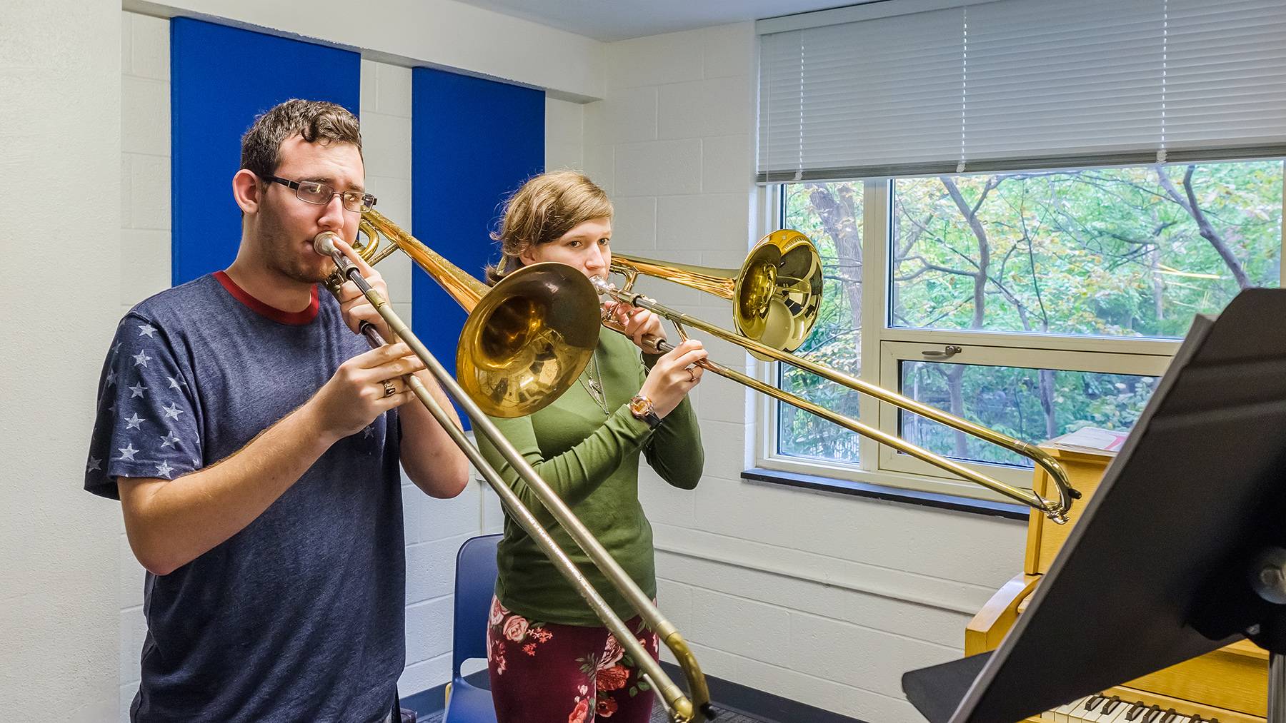 Two UNCG students stand next to a piano while both playing trombones.