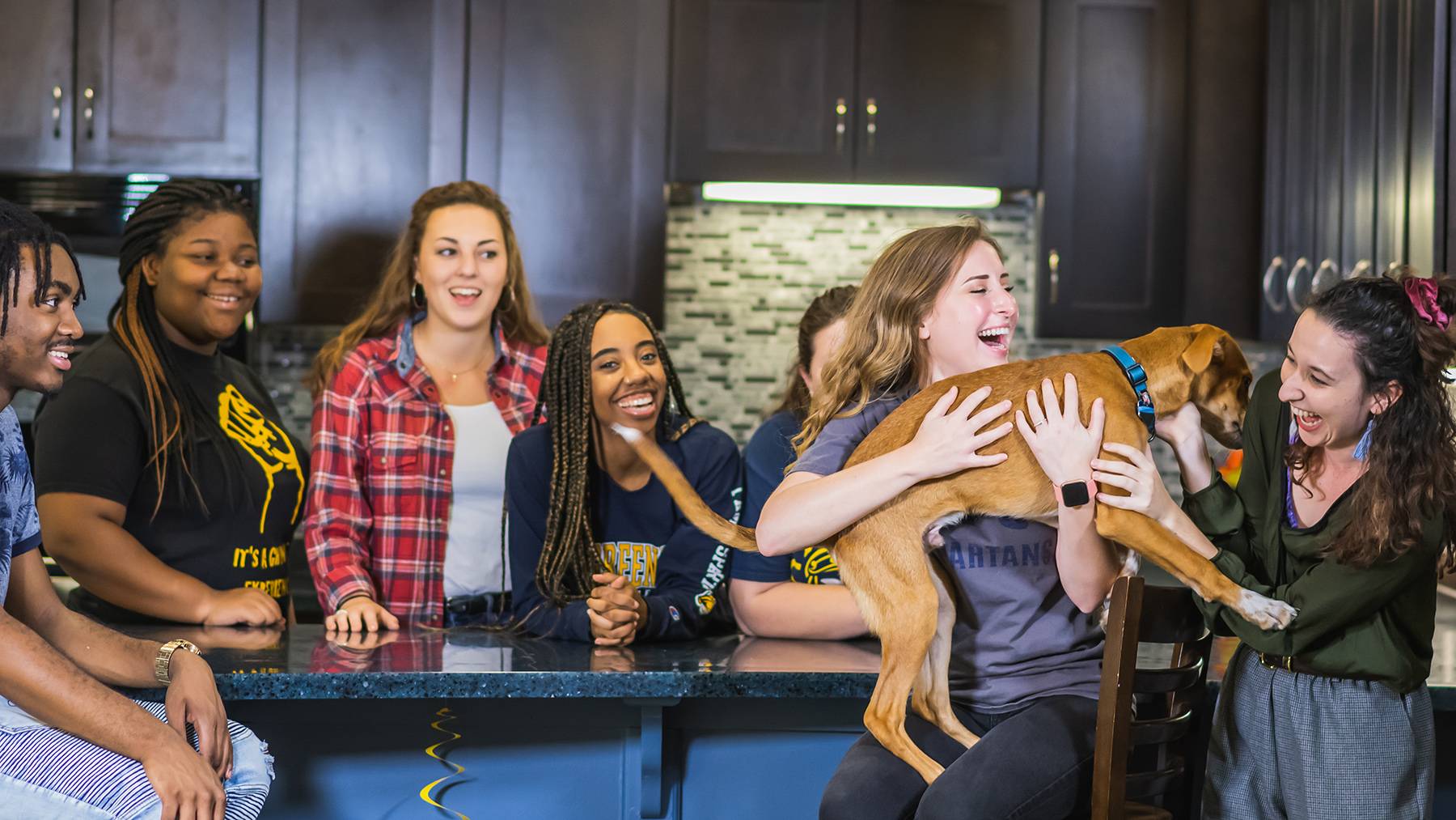 A group of UNCG students in a Spartan Village common room laugh as they play with a puppy.