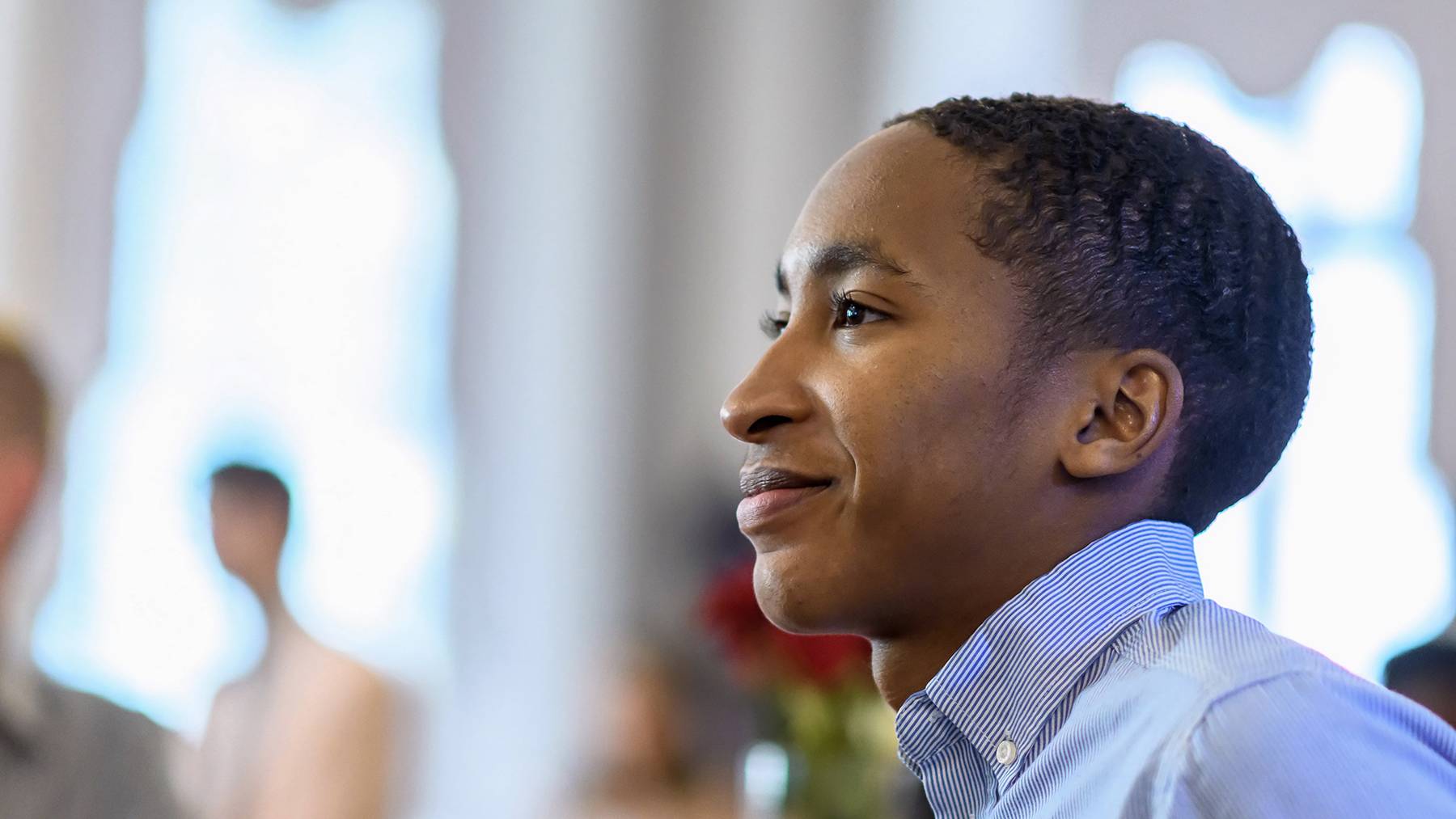Young man smiles as he listens to a speaker during a recognition ceremony.