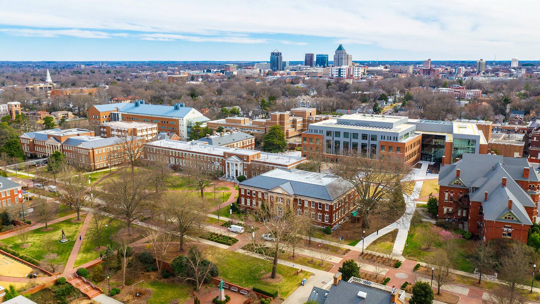 An aerial photo shows the UNCG campus with bare trees during winter, with College Avenue in the foreground and the downtown Greensboro skyline in the background.