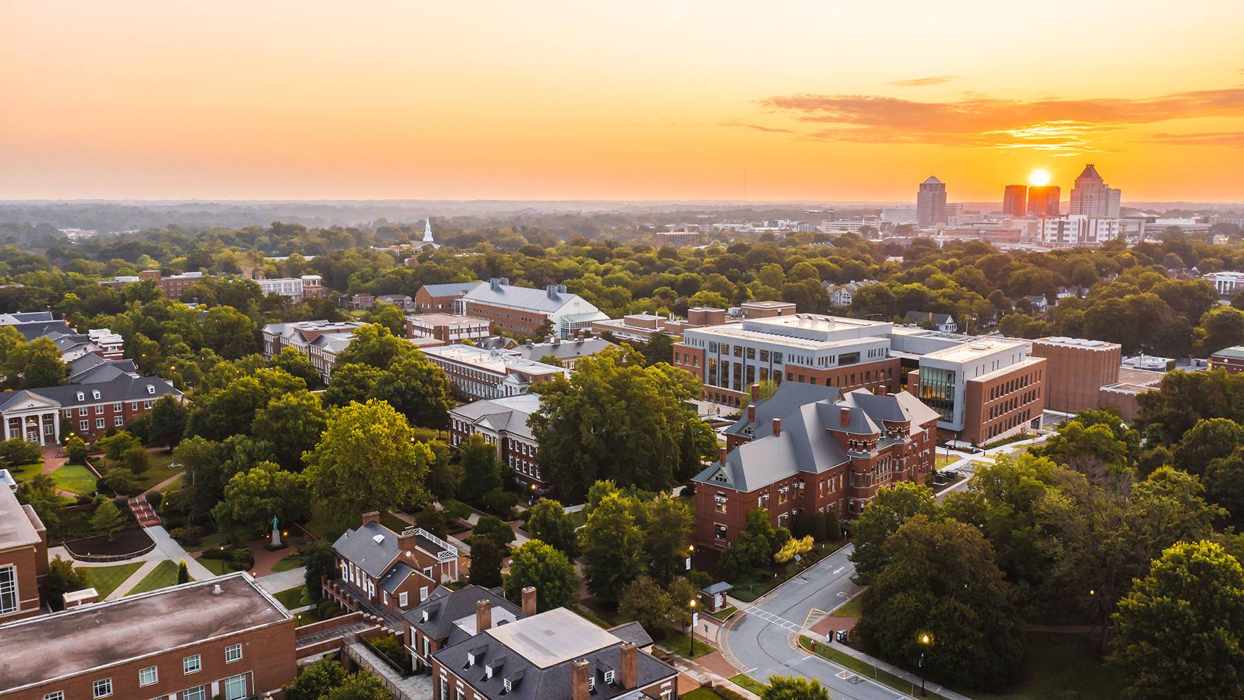 An aerial shot shows the UNCG campus at sunset in the foreground with the downtown Greensboro skyline in the background.