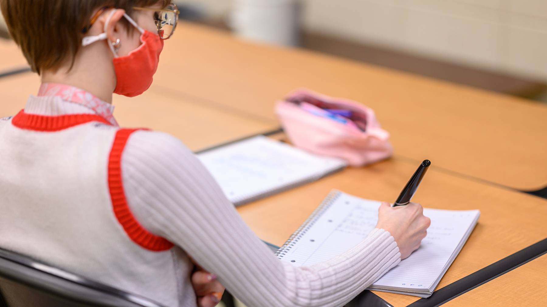 A student wearing a red face mask taking notes while sitting at a classroom desk.