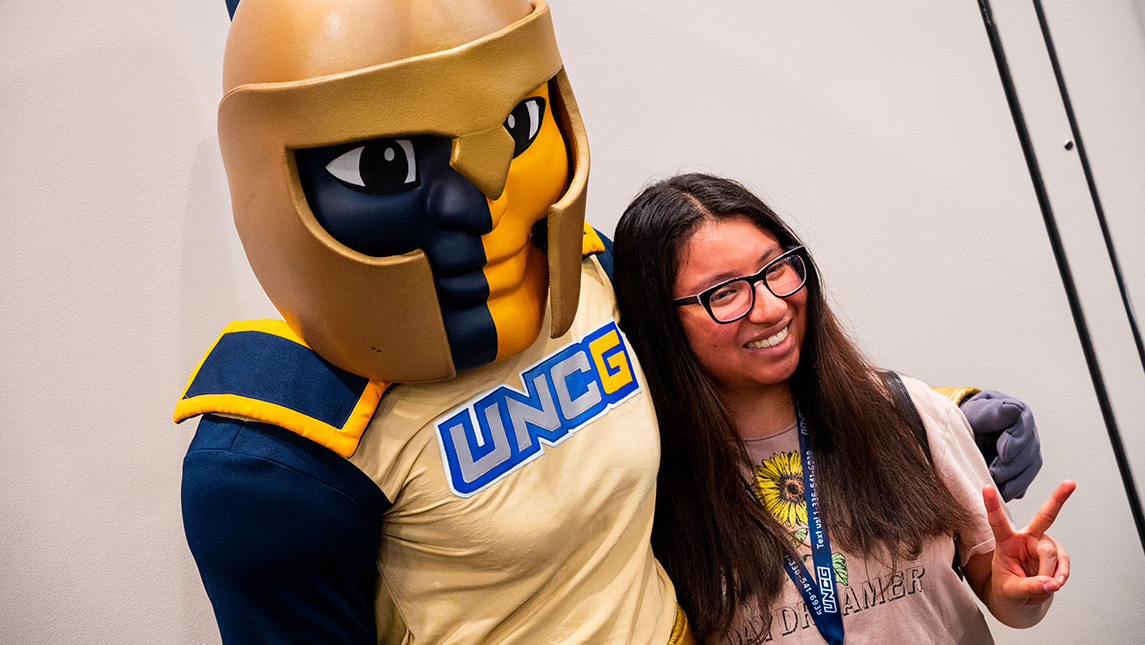 A new student flashes the peace sign while taking a picture with the Spiro mascot.