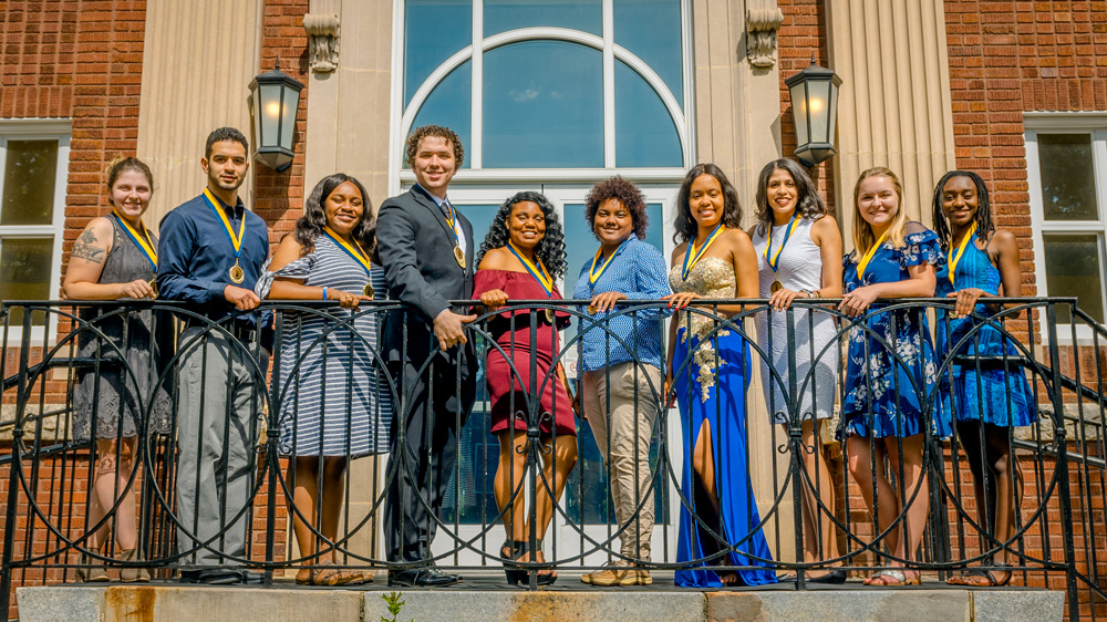 A group of smiling McNair scholars posing on the steps of the Forney Building, each wearing a blue and gold ribbon.