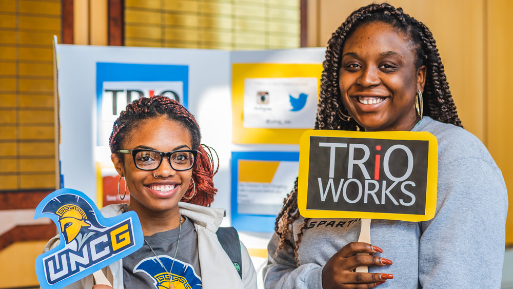 Two smiling students standing in front of a display board. One holds a sign that reads “TRiO Works” the other holds a sign that says, “UNCG” with the spartan head logo.