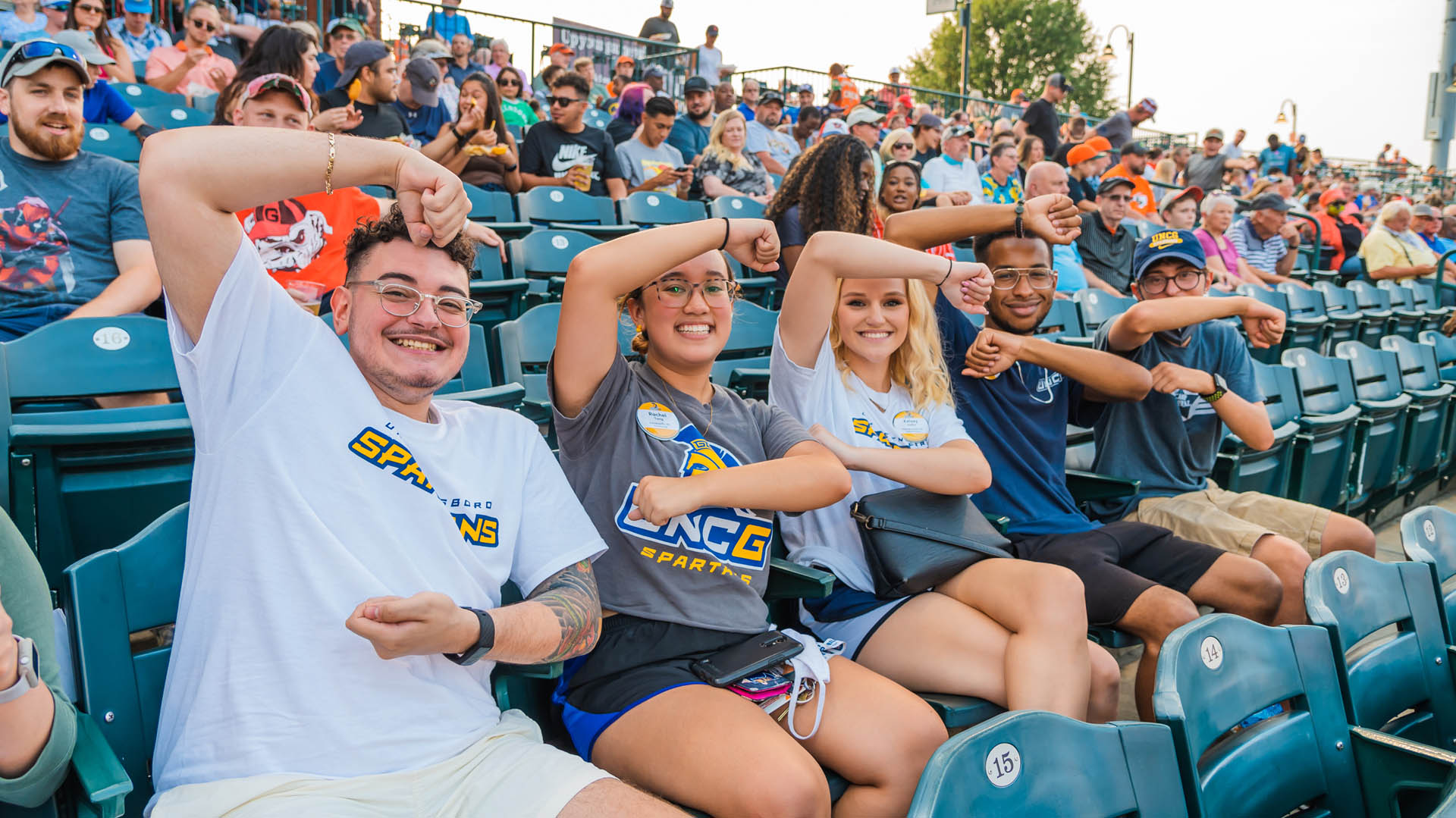 UNCG students in the stands at a Greensboro Grasshoppers game make the “G’ sign with their arms