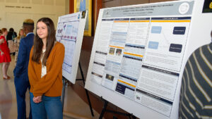 A woman in an orange sweater stands next to her research poster at a conference.