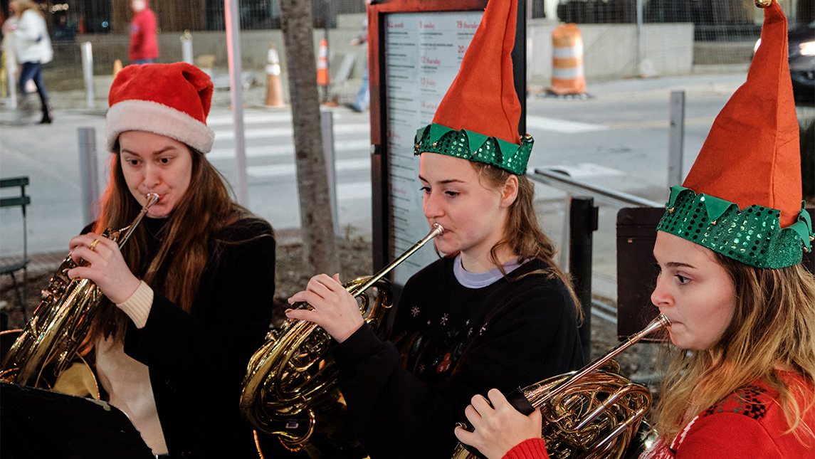 Students in Santa and elf hats play french horns on the street.