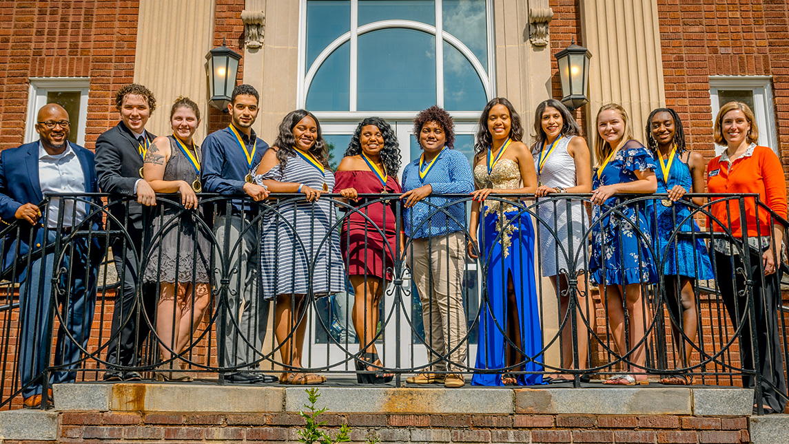 Ten members of the McNair scholar program pose on a balcony with their medals and their advisors upon graduation.