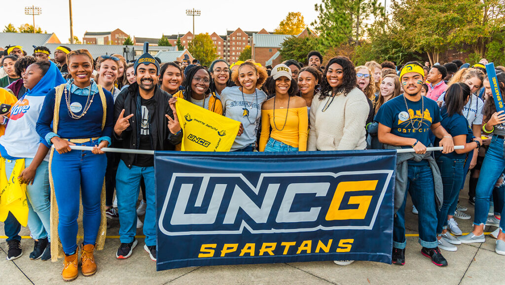 A crowd of students gather around a "UNCG Spartans" banner.