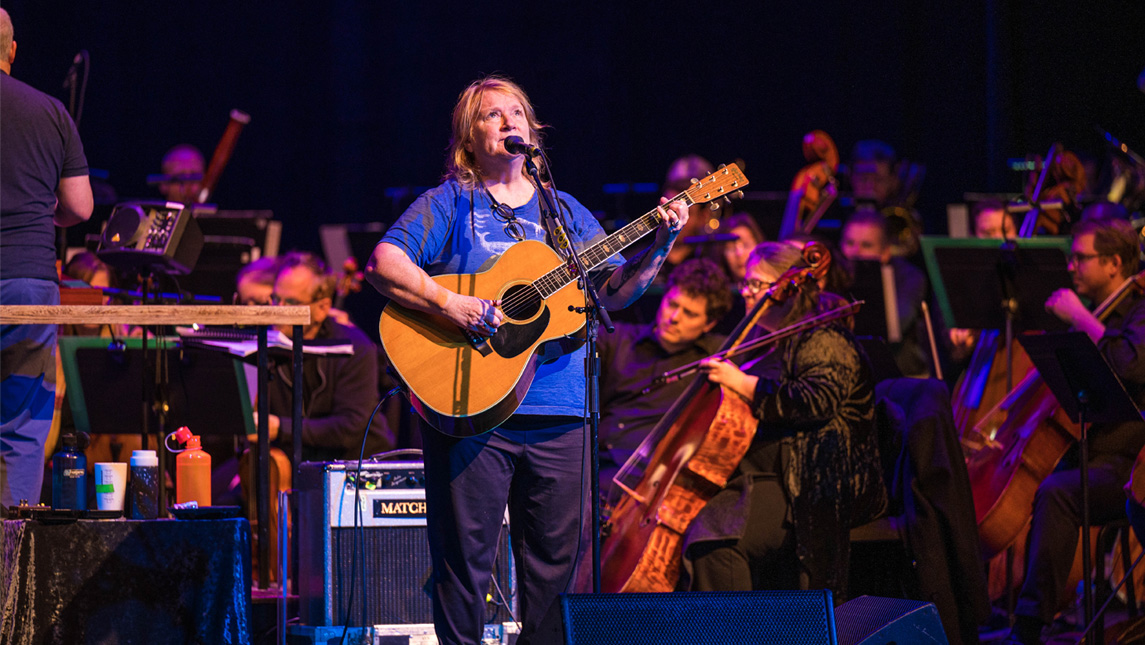 A woman in a blue shirt plays a guitar and sings in front of an orchestra on a stage.