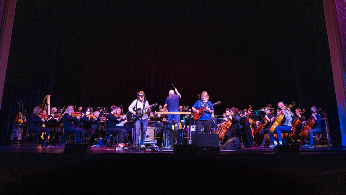 Two women play guitars and sing in front of an orchestra on a stage. The male orchestra conductor has his hand up directing.