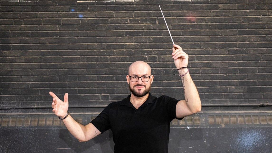Man in black shirt with glasses lifts a conducting baton