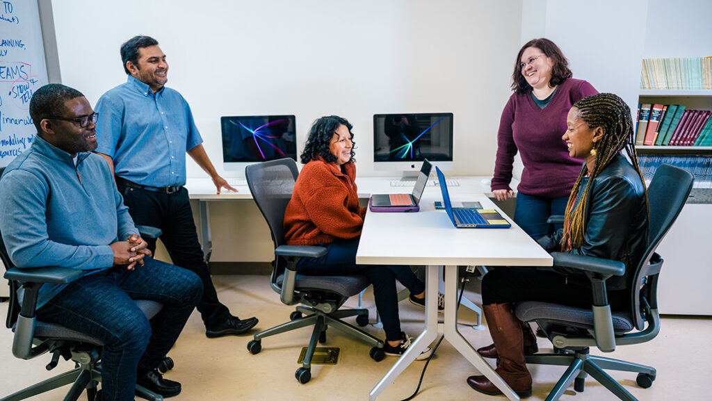 A group of students and a faculty member chat and smile around a table with their computers.