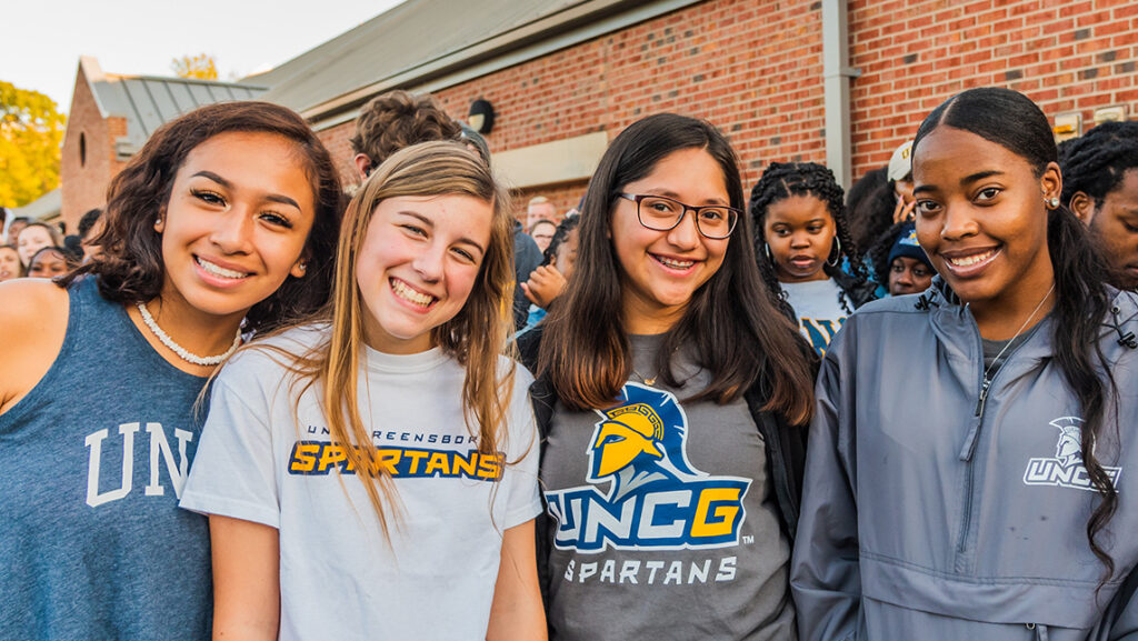 Four female students in UNCG t-shirts smile before the Storm the Streets walk.