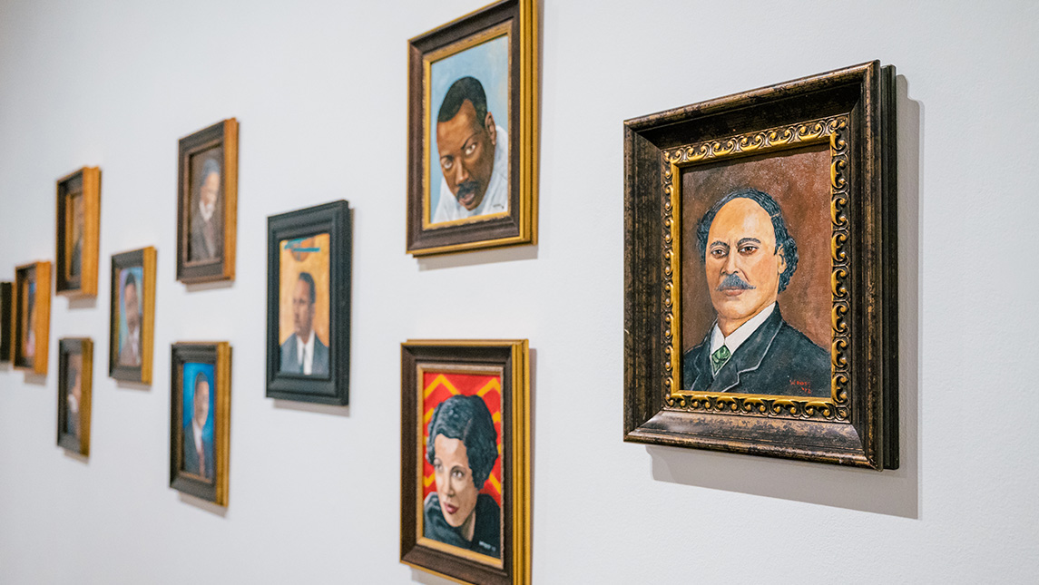 Paintings of headshots of African American artists throughout history in frames hanging on a white wall. 