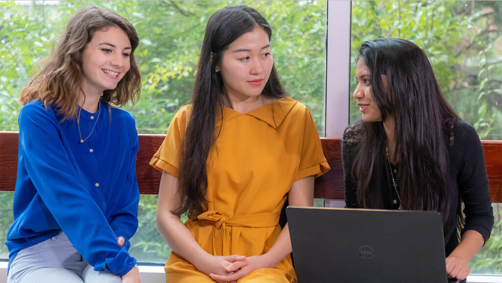 Three female students talk together around a laptop.