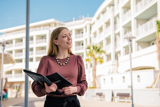 Woman in business attire holds a laptop as she looks into the distance. She stands in front of a white hotel with balconies and palm trees and blue skies overhead.