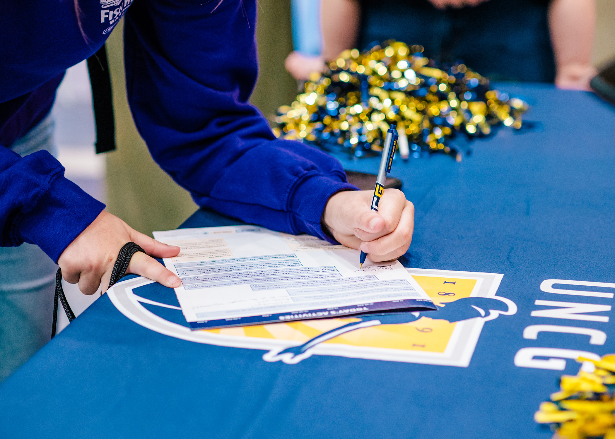 Close-up of person filling out an application with a UNCG pen, on a table with a UNCG tablecloth & pompoms. 
