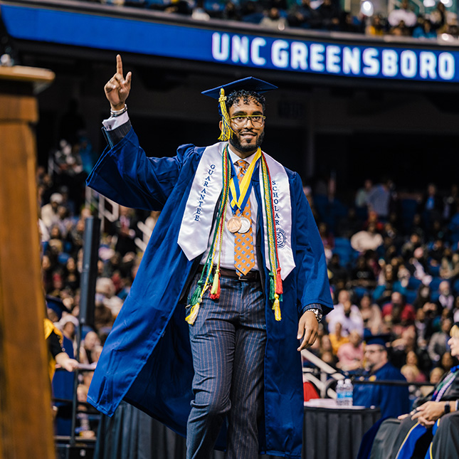 Graduate with lots of honors cords crosses the stage at commencement, pointing up towards a UNCG sign.