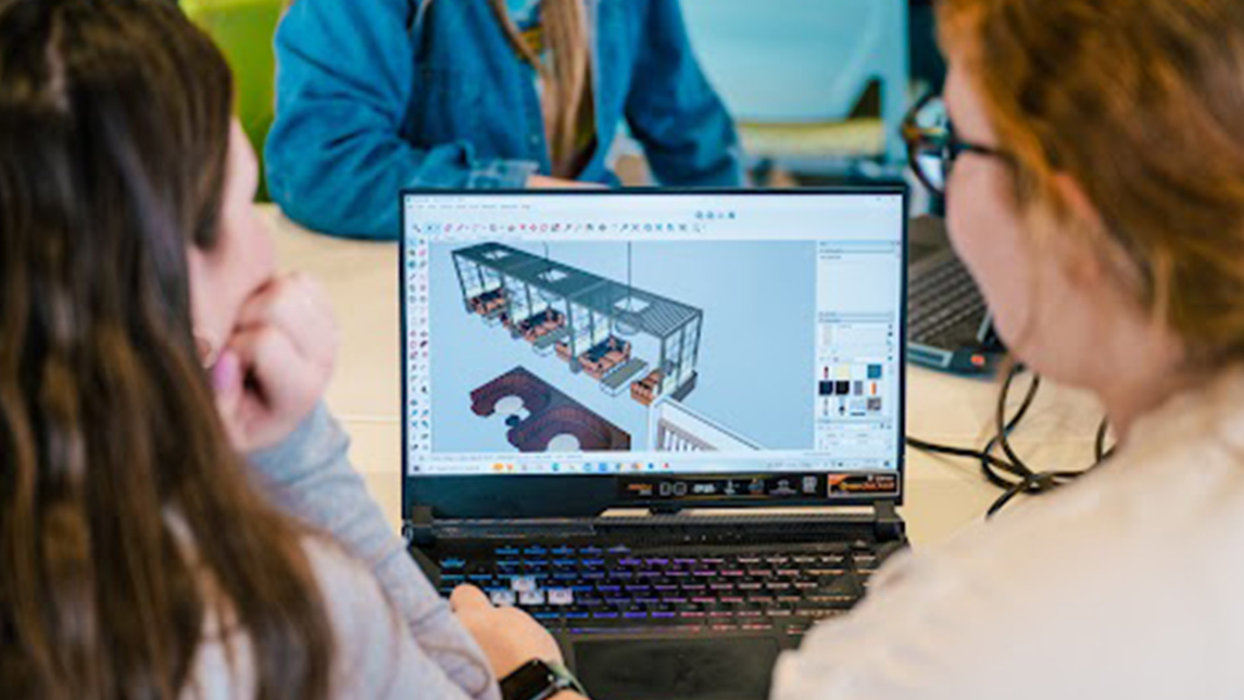Looking over the shoulders of two students who are working on a laptop that features 3-D models of a restaurant's interior.