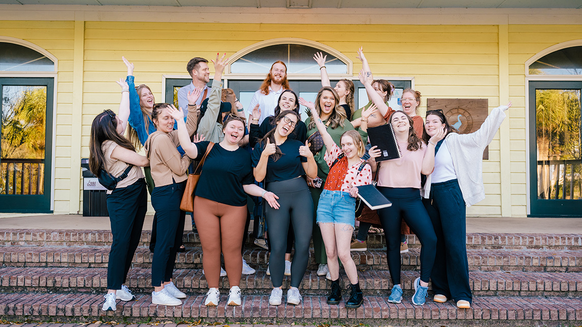 16 students and a teacher in a crazy group pose on a porch with notebooks