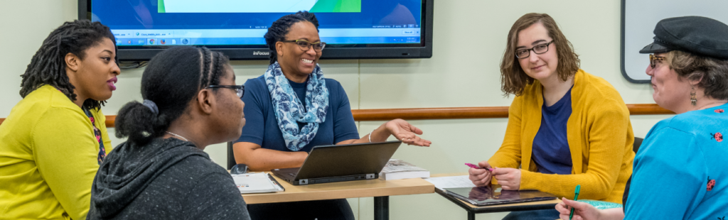 Seated behind desks, LIS students engage in a group discussion. In the background, a large screen displays a breakdown of multijurisdictional libraries in North Carolina.