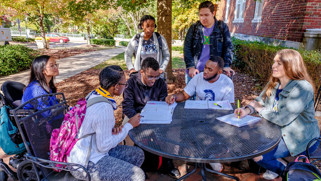 A group of students sit or stand around a table working on homework together.