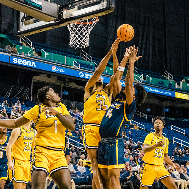 UNCG and Towson basketball players jump for the ball under the basket.