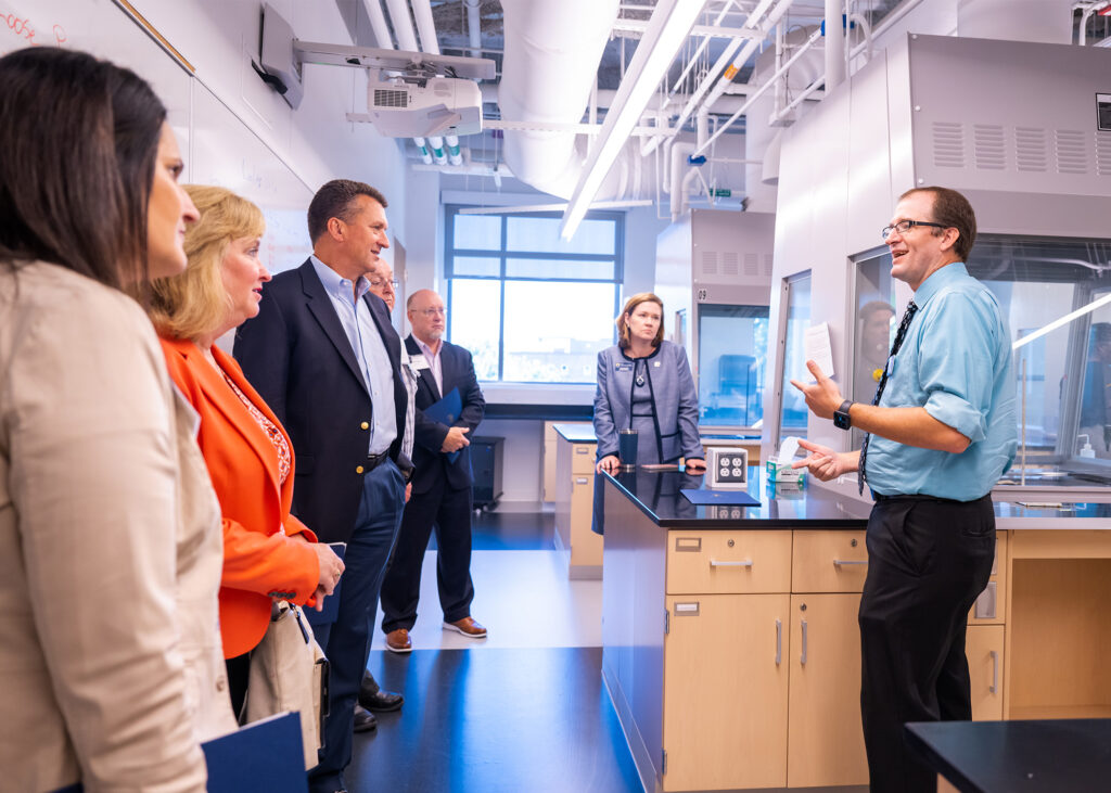 People dressed in business attire listen to a man speak in a lab.