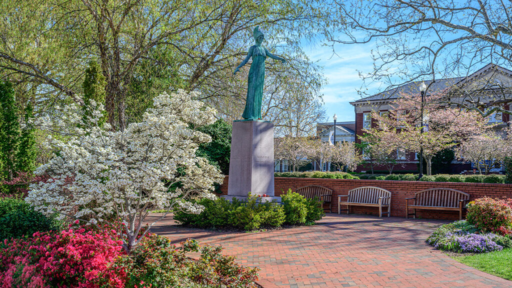 Statue of Minerva in springtime with dogwoods and azaleas blooming around it and campus buildings in background.